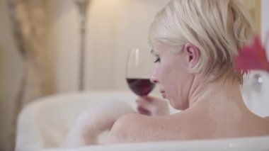 Banyo köpüğüyle küvette oturan ve elinde kırmızı şarapla bardak tutan genç sarışın kadının yakın çekimi. Kafkasyalı güzel bir bayanın alkolle banyo yaptığı arka manzara. Omzundan vuruluyor. Neşe, yaşam tarzı.