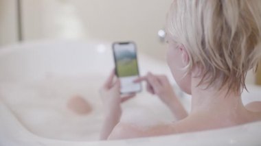 Sarışın kadın banyo yaparken sosyal medyayı karıştırıyor. Küvette köpükle akıllı telefonun bulanık ekranına bakan beyaz bir kadının arka görüntüsü. İnternet sörfü, yaşam tarzı, bağımlılık.