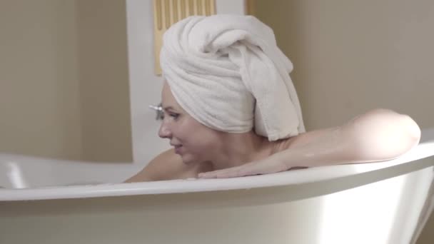 Glada vuxna kvinna tar badskum och blåser det till kameran. Porträtt av glad ung vit dam i hår handduk har kul i badkar. Lycka, glädje, fritid, livsstil, avkoppling. — Stockvideo