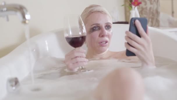 Femme blonde pleurant lisant des messages sur smartphone et buvant du vin rouge. Portrait de dame caucasienne déprimée avec un mascara coulant dans une baignoire mousseuse. Hystérique, dépression, problème, mode de vie . — Video