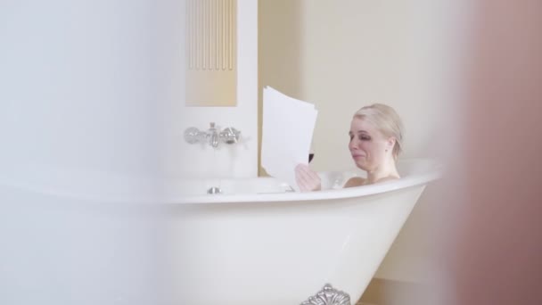 Depressed woman crying in bathtub and drinking wine. Side view of stressed blond Caucasian lady reading documents and weeping. Problems, troubles, stress, depression, despair. Shooting through doorway — Stock Video