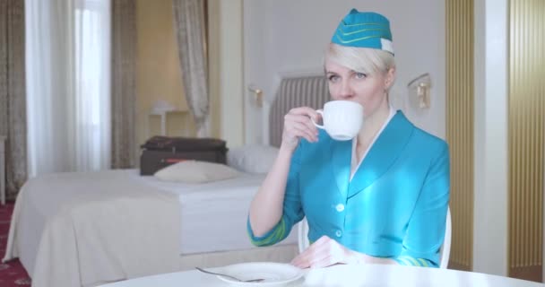 Retrato de la encantadora azafata caucásica en uniforme azul bebiendo café por la mañana en la habitación del hotel. Mujer rubia confiada descansando antes del vuelo. Elegancia, estilo de vida. Sede del cine 4k ProRes . — Vídeos de Stock