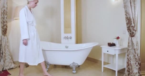 하얀 목욕 복을 입은 멋진 백인 여성 이 호화 로운 욕실로 들어가 욕조에 앉아 있습니다. 다리를 가로지르는 금발 여자의 넓은 샷. 목욕, 생활 방식, 위생. 시네마 4k ProRes HQ. — 비디오