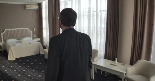 Vista trasera de hombre caucásico confiado en chaqueta formal y anteojos mirando alrededor de la habitación del hotel. Elegante hombre de negocios senior que llega en alojamiento durante su viaje de negocios. Sede del cine 4k ProRes . — Vídeos de Stock