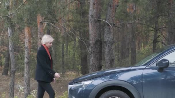 一个自信的金发女人打开车篷,看着发动机的侧视图.成年女司机站在森林旁边的破车旁边。农村道路上的车祸。生活方式. — 图库视频影像