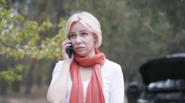 Gergin, sarışın, beyaz bir kadın telefonda konuşuyor ve arka planda bozuk bir arabayla el kol hareketi yapıyor. Stresli kadın sürücünün portresi banliyö yolunda sigorta istiyor.