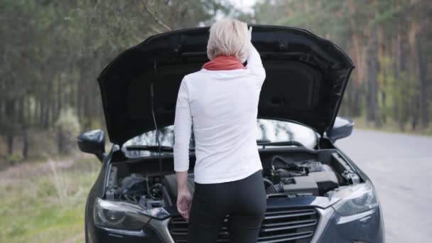 Widok z tyłu szczupłej białej blondynki kładącej ręce na biodrach na tle zepsutego samochodu. Stresowana kobieta kierowcy stojąca przed otwartym kapturem i patrząc na silnik. Pęknięcie, problem. — Wideo stockowe