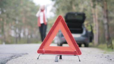 Bulanık beyaz kadın, ön planda duran acil dur levhasıyla telefonda konuşuyor. Kadın sürücü banliyö yolundaki araba arızasıyla ilgili sorun yaşıyor. Stres, kaza, bela.