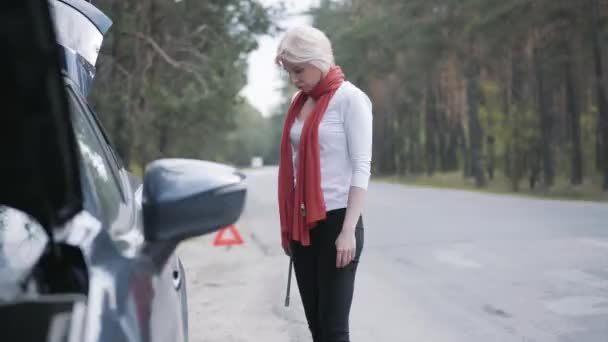 Sidovy av vuxen vit kvinna med skiftnyckel åtdragning skruvar av bil hjul utomhus. Självsäker blond elegant dam laga bil brott själv på förortsväg. Reseproblem. — Stockvideo