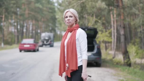 Widok z boku dorosłej blondynki stojącej obok zepsutego samochodu z kluczem i czekającej na lawetę. Portret smutnej białoruskiej kobiety kierującej pojazdem po wypadku drogowym na wsi. — Wideo stockowe