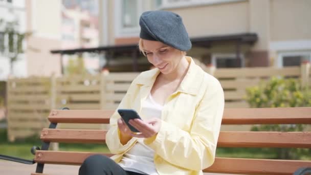 一个快乐优雅的高加索女人用智能手机打字，面带微笑的画像。快乐的金发女士穿着黄色外套，戴着帽子在网上发短信。休闲、欢乐、生活方式、无线通信. — 图库视频影像