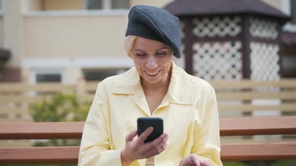 Mujer joven sonriente mirando la pantalla del teléfono inteligente y riendo en voz alta. Retrato de una elegante dama rubia caucásica sentada en el banco y usando las redes sociales al aire libre. Estilo de vida, felicidad . — Vídeos de Stock