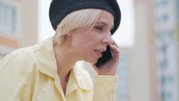 Роздратована красива жінка сперечається по телефону на вулиці. Портрет роздратованої кавказької жінки в елегантному капелюсі і жовтому пальто свариться на вулицях міста. Спосіб життя, стрес. — стокове відео