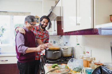 Mid adult man is looking over his father's shoulder as he prepares a curry at home.  clipart