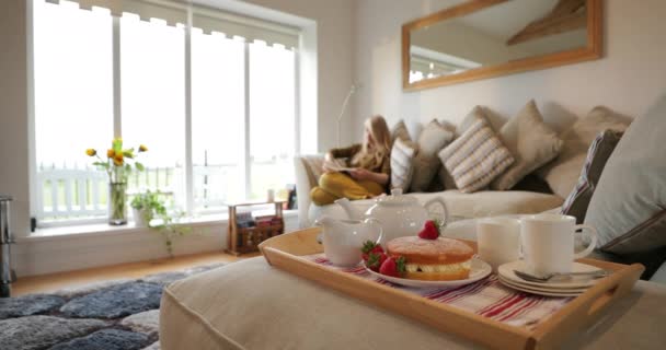 在客厅的托盘上挂着一张奶油蛋糕和茶具的特写照片 可以看到一个女人在沙发后面看书 — 图库视频影像