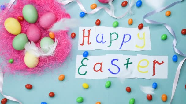 青い背景にピンクの巣の中のイースターカラーの卵 テーブルの上のキャンディとお菓子 テキスト Happy Easter — ストック動画