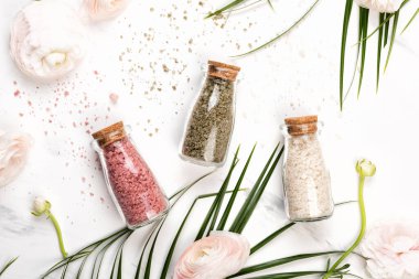 Cam banyo tuzu şişeleri. Pastel pembe ranunculus çiçekleri ve beyaz mermer zemin üzerinde palmiye yaprakları düz ya da üst görünüm. Kozmetik kaplıca konsepti