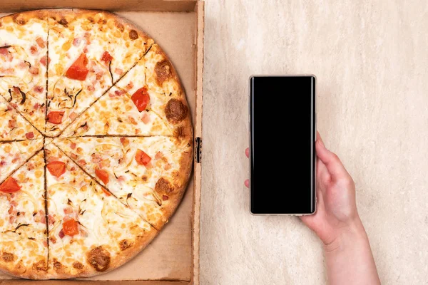 Pizza in a cardboard box and phone in hand on a light marble background. Copy space for your text. Top view, flat lay. Takeaway food concept
