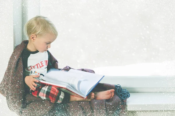 Çocuk pencerenin yanında kitap okuyor. Kar yağarken güzel kış havası — Stok fotoğraf