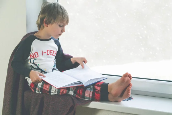 Çocuk pencerenin yanında kitap okuyor. Kar yağarken güzel kış havası — Stok fotoğraf