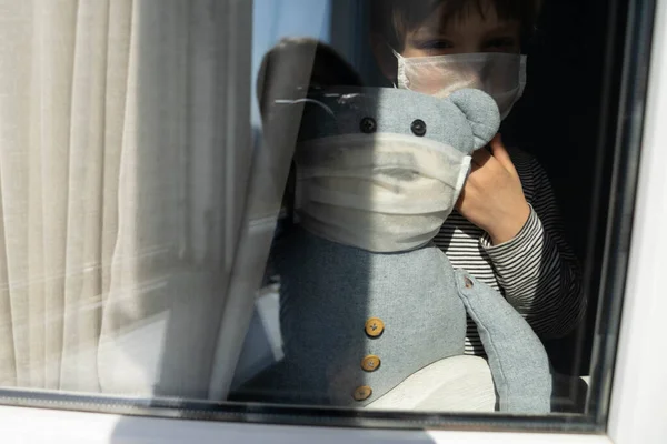 Коронавирусная концепция - карантин, социальное дистанцирование, мальчик в медицинской маске в окне — стоковое фото