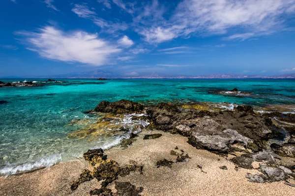 A costa da ilha de Chrissy em um dia ensolarado de verão com água do mar azul-turquesa, um fundo rochoso, pedras pretas em primeiro plano e um céu azul nublado com neblina. Creta, Grécia . — Fotografia de Stock