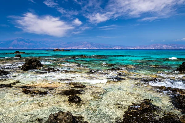 The coast of Chrissy island on a sunny summer day with turquoise sea water, a rocky bottom, black stones in the foreground and a blue cloudy sky with haze. Crete, Greece. — Stock Photo, Image