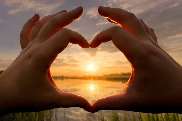 Mains en forme de cœur contre le coucher du soleil sur le lac ou la rivière en été avec un fond nuageux ciel. Paysage . — Photo