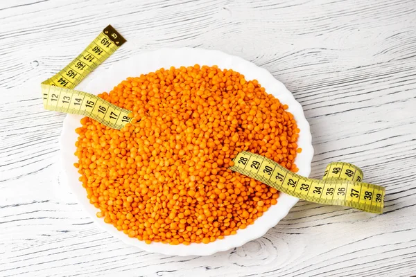 Grains of red lentils and a measuring tape in a plate on a white wooden table. The concept of weight loss and proper nutrition.