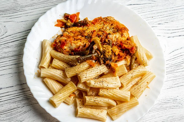 Cooked Italian tortiglioni pasta with turkey steak chop under vegetables and tomato paste in a plate on a white wooden table. Traditional cuisine.