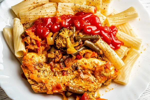 Cooked Italian tortiglioni pasta with turkey steak chop under vegetables and tomato paste in a plate on a white wooden table. Traditional cuisine.