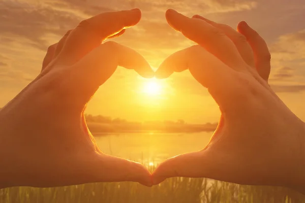 Hands in the shape of heart against the sunset over the lake or river in the summer with a cloudy sky background. Landscape.