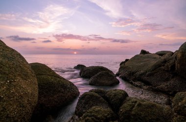 Dramatik deniz manzarası, ön planda kayalar, gün batımı manzarası arka plan.