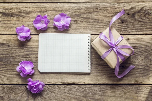 Background with violet flowers, gift box and empty notebook for — Stock Photo, Image