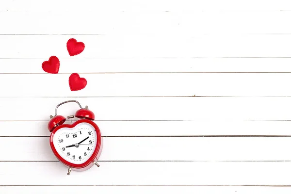 Heart-shaped alarm clock and hearts on white woooden background.