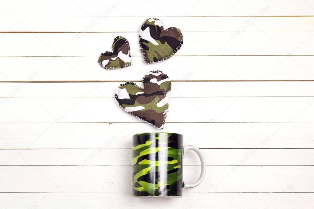 Camouflage mug with khaki hearts on a white wooden background. C