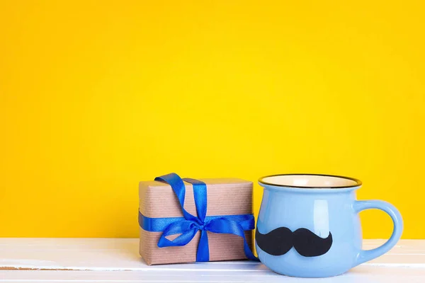 Blue mug with a mustache and gift box on yellow background. Copy