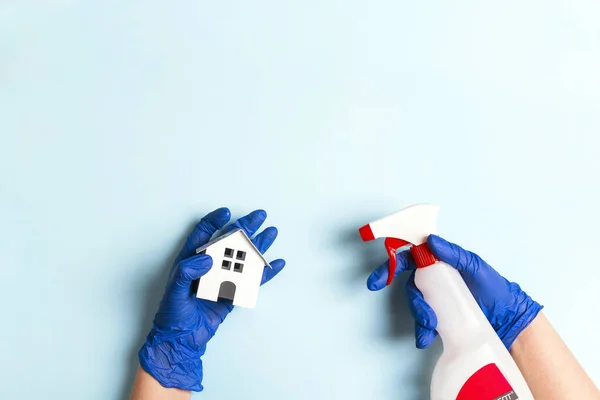 Female hands in rubber gloves disinfect a toy house with an alcohol spray for coronavirus protection. House disinfection concept. Blue background with copy space for text.