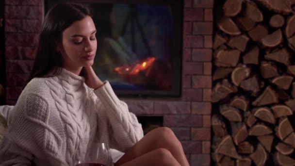 若い女性が自宅で暖炉のそばで座って、赤ワインを飲む. — ストック動画