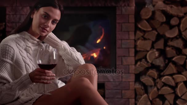 Mujer joven sentada en casa junto a la chimenea y bebiendo un vino tinto . — Vídeos de Stock