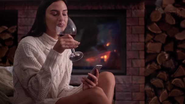 Молода жінка сидить вдома біля каміна і п'є червоне вино . — стокове відео
