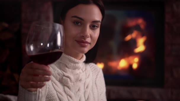 若い女性が自宅で暖炉のそばで座って、赤ワインを飲む. — ストック動画