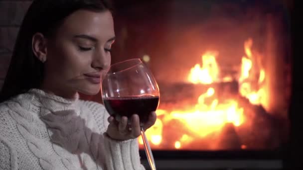 Mujer joven sentada en casa junto a la chimenea y bebiendo un vino tinto . — Vídeos de Stock