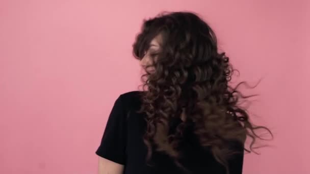 Menina na moda bonita com cabelo encaracolado longo em uma camiseta preta. Menina no estúdio em um fundo rosa. Publicidade, produtos capilares, salão de beleza, cosméticos, roupas. Moda, boutique. Rosa . — Vídeo de Stock