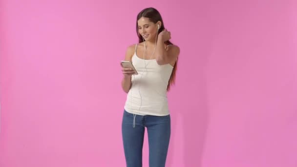 En tjej i jeans och en T-shirt som lyssnar på musik i hörlurar på en rosa bakgrund — Stockvideo