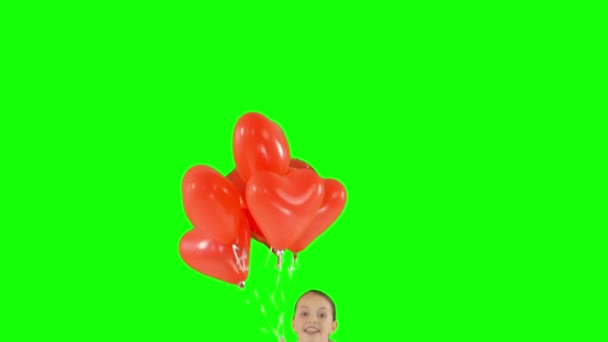 Маленькая девочка держит связку воздушных шаров в форме красного сердца. Выпрыгивает. изолированный на зеленом экране. Стрельба медленным движением — стоковое видео