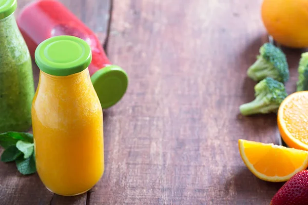 Frische Erdbeer-, Orangen- und Brokkoli-Smoothie in Flaschen mit grünen Verschlüssen mit Obst und Gemüse auf braunem rustikalem Holzhintergrund, Kopierraum in der Mitte — Stockfoto