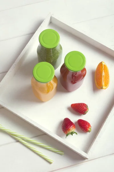 Fresh orange, strawberry and broccoli smoothie in bottles with green caps with fruits and mint in a white wooden rustic box and on a white background, vertical.