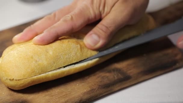 Man cuts bread h in a half by sharp knife on a wooden board at the kitchen, making sandwich with spanish jamon — 비디오