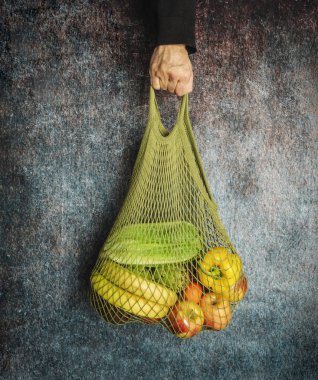 A man's hand holds a green string bag with fresh vegetables and 